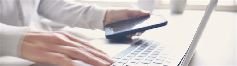 En person sitter vid en bärbar dator och skriver med ena handen och håller en smartphone i andra handen