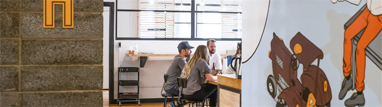 Kontorsarbetare i ett mötesrum i öppet landskap sedda från en korridor med lekfulla väggmålningar..