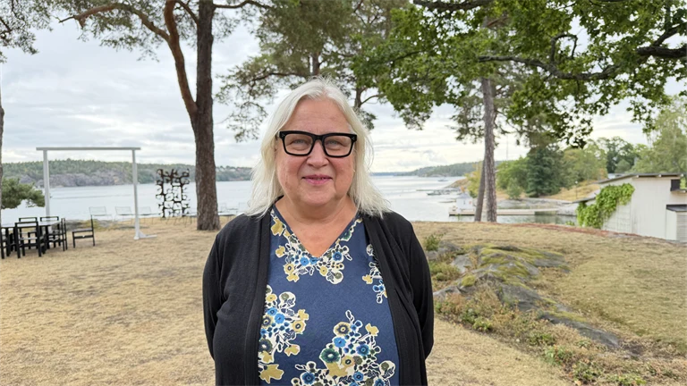 Forskare Maria Åkesson har ljust hår, glasögon med mörka bågar, en mellanblå blus med blommiga mönster och en mörkblå kofta, i bakgrunden syns utsikt över vatten och grönområden.