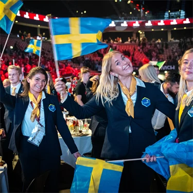 Några av ungdomarna i yrkeslandslaget ler och viftar med svenska flaggor vid middagsbordet under invigningen av Yrkes-EM.