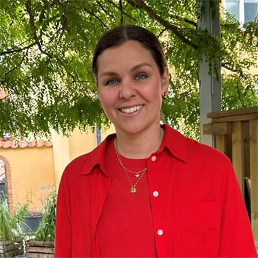 Lina Ejlertsson står under ett lövträd och ser in i kameran. Hon har på sig en röd tröja, röd skjorta och vita byxor.