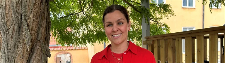 Lina Ejlertsson står under ett lövträd och ser in i kameran. Hon har på sig en röd tröja, röd skjorta och vita byxor.