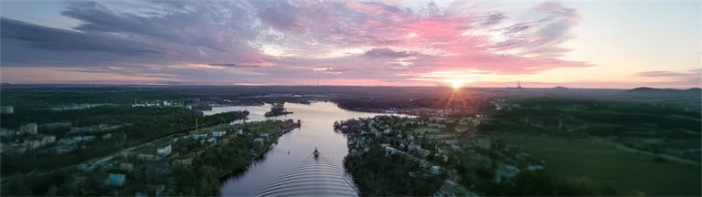 En flod i en fjärd omgiven av skog sedd uppifrån, i solnedgången, med rosa och lila moln på ljus himmel. Redigerad med AI: