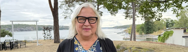 Forskare Maria Åkesson har ljust hår, glasögon med mörka bågar, en mellanblå blus med blommiga mönster och en mörkblå kofta, i bakgrunden syns utsikt över vatten och grönområden.