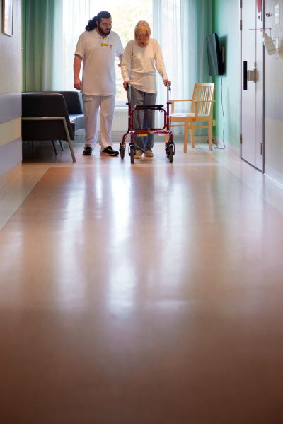 Vårdare går bredvid kvinna med rollator i sjukhuskorridor