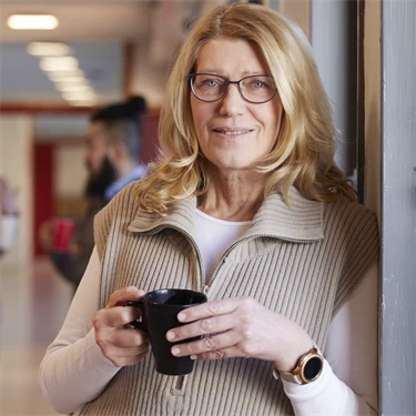 kvinna som står i skolkorridor med en kaffekopp i handen och ler mot kameran