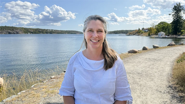 Forskare Eva Lindell har en vit blus och ler mot kameran, i bakgrunden syns utsikt över vatten och grönområden.