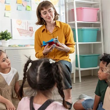 En förskolepedagog står framför en barngrupp. Pedagogen ler och håller upp ett flygplan, ett barn räcker upp handen. 