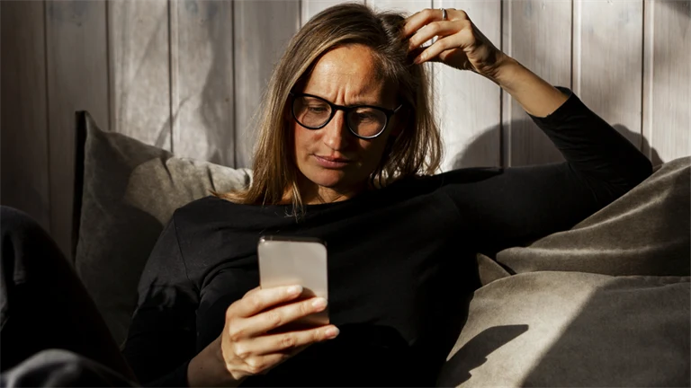 Kvinna sitter i soffan och tittar på sin mobiltelefon med bekymrad min.