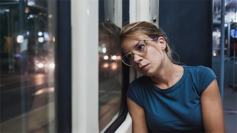 Kvinna med glasögon och blå t-shirt sitter på spårvagn och lutar huvudet mot fönstret