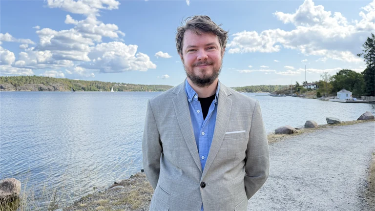 Forskare Andreas Bergqvist har skägg, ljusblå skjorta och ljusgrå kavaj, i bakgrunden syns utsikt över vatten och grönområden.