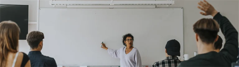 Lärare står och pekar på whiteboard i klassrum med elever i övre tonåren