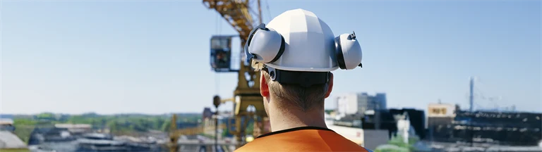 Manlig byggnadsarbetare med hjälm och hörselskydd blickar ut över lyftkran