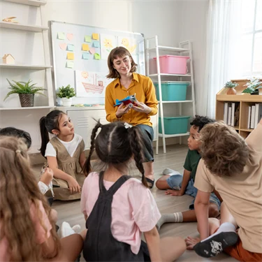 En förskolepedagog står framför en barngrupp. Pedagogen ler och håller upp ett flygplan, ett barn räcker upp handen. 