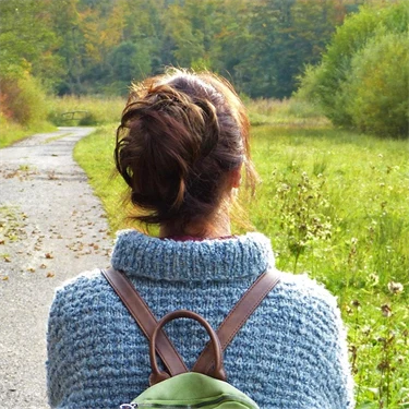 Kvinna med uppsatt hår och ryggsäck går på en stig mot en höstfärgad skog.