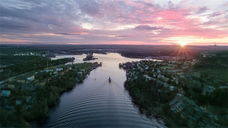 En flod i en fjärd omgiven av skog sedd uppifrån, i solnedgången, med rosa och lila moln på ljus himmel. Redigerad med AI: