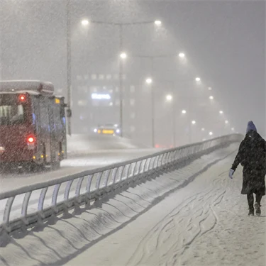 Snöig väg där en röd buss passerar en kvinna som går på trottoaren. 