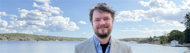 Forskare Andreas Bergqvist har skägg, ljusblå skjorta och ljusgrå kavaj, i bakgrunden syns utsikt över vatten och grönområden.