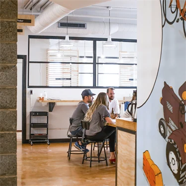 Kontorsarbetare i ett mötesrum i öppet landskap sedda från en korridor med lekfulla väggmålningar..