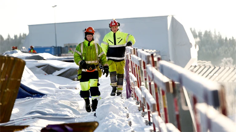 Personer i varselkläder och hjälm jobbar utomhus i ett snöigt vinterlandskap.