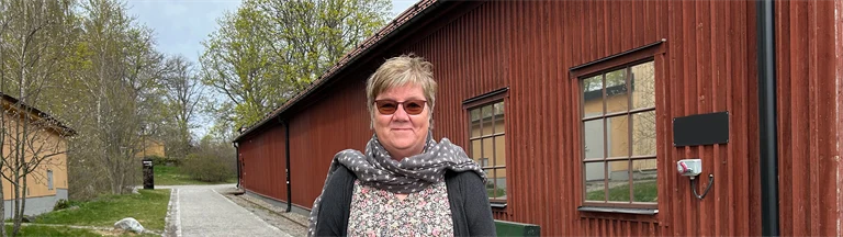 Gunnel Hensing forskare vid Göteborgs universitet står framför ett rödmålat trähus, hon har glasögon, ljusblå jeans, blommig tröja, scarf och en grå lång kofta på sig